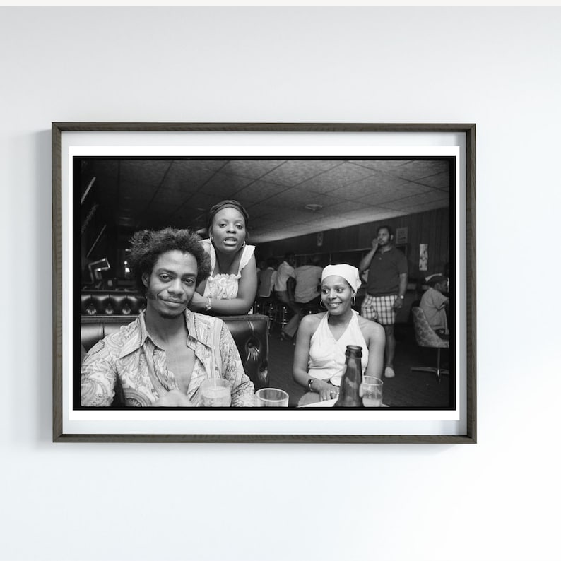 May include: Black and white photo of three people sitting at a bar. The person in the foreground is wearing a patterned shirt and looking at the camera. The person behind them is wearing a white shirt and looking at the camera. The person in the background is wearing a white hat and looking at the camera.