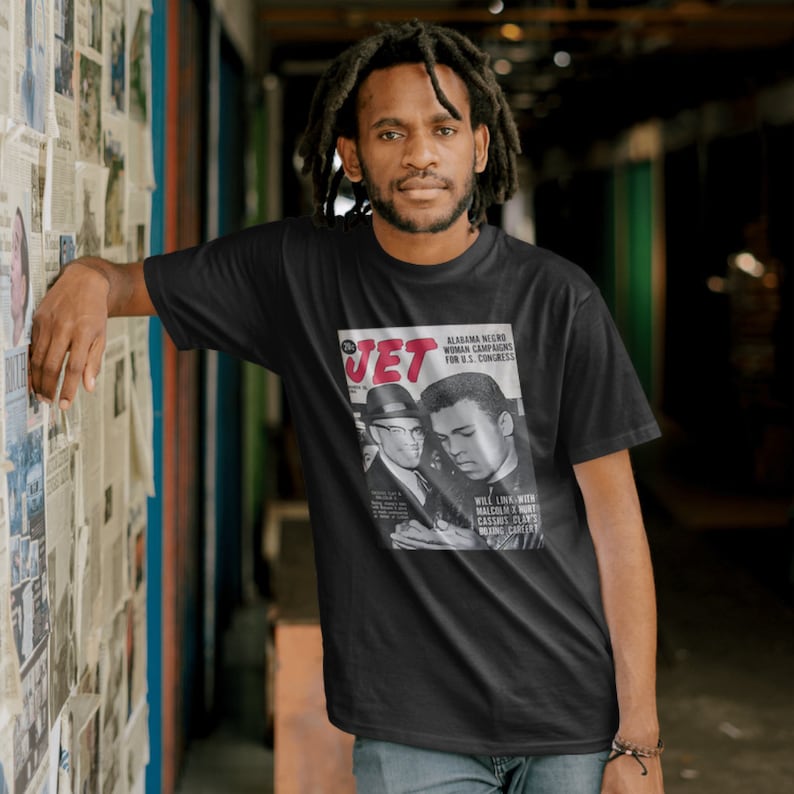 May include: A black t-shirt with a vintage Jet magazine cover featuring a photo of Muhammad Ali and Malcolm X. The magazine cover text reads "Alabama Negro Woman Campaigns for U.S. Congress" and "Will Link With Malcolm X, Wurt Cassius Clay's Boxing Cartert".