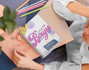 Pode incluir: Uma agenda espiralada rosa e branca com o texto "The Bougie Lifestyle Planner" na capa. Um passaporte é visível na capa.