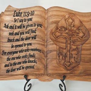 May include: Wooden book-shaped plaque with engraved text from Luke 11:9-10 and a cross design. The plaque is supported by a black metal stand. The text is in black, and the wood has a natural grain.