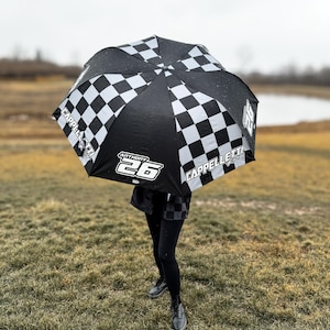 Op de afbeelding: Een zwart-wit geblokte paraplu met de naam "CAPPELLETTI" en het nummer "26" erop gedrukt. De paraplu wordt vastgehouden door een persoon die een zwarte legging en zwarte schoenen draagt.