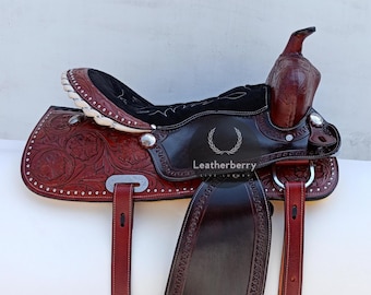 May include: A brown leather saddle with intricate floral tooling and silver accents. The saddle features a black suede seat and a black leather skirt. The saddle is accompanied by two stirrups and a cinch.