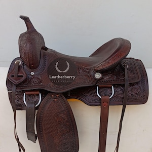 Puede incluir: Una silla de montar de cuero marrón con intrincados detalles florales y acentos plateados. La silla de montar tiene el logotipo "Leatherberry" y está diseñada para montar a caballo.