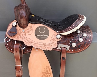 May include: A brown leather saddle with tooled floral designs and silver accents. The saddle has a black seat and a brown horn. The saddle is made by Leatherberry.