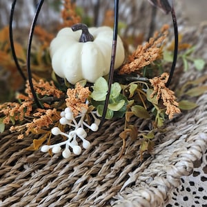 May include: A woven basket centerpiece featuring a black metal pumpkin frame, a small white decorative pumpkin, artificial orange foliage, green leaves, and white berries. The arrangement is set within a textured, natural-toned woven basket.