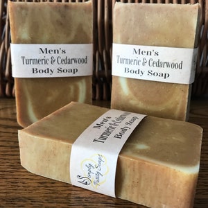 Peut inclure: Trois savons avec une étiquette indiquant "Savon pour le corps au curcuma et au cèdre pour homme". Le savon est de couleur brun clair avec des tourbillons blancs.