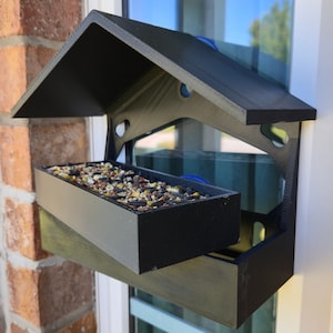 May include: A black bird feeder with a roof and a tray for birdseed. The feeder is mounted on a window.