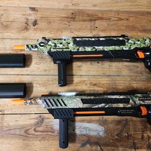 May include: Two black and green camouflage toy guns with orange accents. The guns have a black stock and a black barrel. The guns are on a wooden surface. The text on the gun reads "BUG-A-SALT", "MADE IN CHINA", and "RACE TO EXTER",