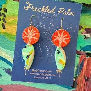 May include: A pair of handmade earrings. Each earring has a round orange top with a white tree design, and a teardrop-shaped bottom in blue, green, and yellow. The earrings are displayed on a blue card with the text "Freckled Palm".