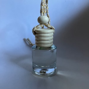 Puede incluir: Una botella de vidrio transparente con una tapa de madera blanca y una cuerda dorada y blanca colgando de ella. La botella está llena de un líquido transparente.