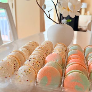 May include: A clear plastic tray filled with colorful macarons. Some are white with sprinkles, others are pastel orange and green. A white vase with white flowers is in the background.