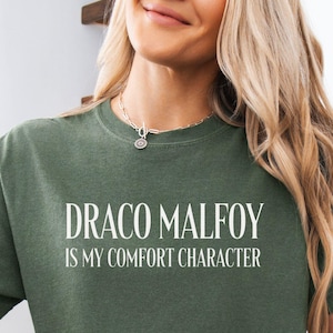 May include: Olive green t-shirt with the text "DRACO MALFOY is my comfort character" in white. The shirt is paired with denim shorts and a beige cardigan. A round sticker with "COMFORT COLORS MOSS" is visible.