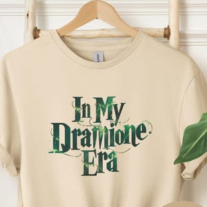 May include: Sand-colored t-shirt with the text "In My Dramione Era" in green and white. The shirt is knotted at the bottom. A wooden hanger and a woven basket are visible in the background.