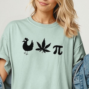 May include: A light green short-sleeve t-shirt featuring a black graphic print of a chicken, a cannabis leaf, and the mathematical pi symbol. The shirt has a crew neckline and a loose fit, layered under a blue denim jacket.