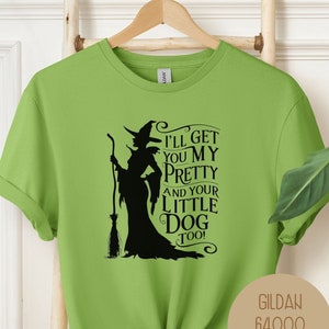 以下が含まれることがあります： ライムグリーンの半袖Tシャツ。ほうきを持った魔女の黒いシルエットのグラフィックが描かれています。テキストは「I'LL GET YOU MY PRETTY AND YOUR LITTLE DOG TOO!」と書かれています。右側の円形のベージュのラベルには「GILDAN 64000 KIWI」と記載されています。