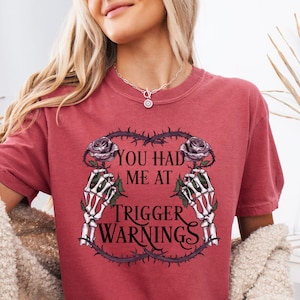 Può includere: T-shirt color cremisi con la scritta "YOU HAD ME AT TRIGGER WARNINGS" in stile gotico, incorniciata da rose e mani di scheletro. La maglietta fa parte della collezione Comfort Colors.