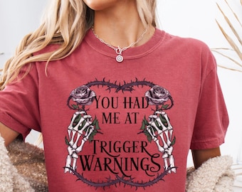 May include: Crimson-colored t-shirt with the text "YOU HAD ME AT TRIGGER WARNINGS" in a gothic font, framed by roses and skeleton hands. The shirt is part of the Comfort Colors collection.