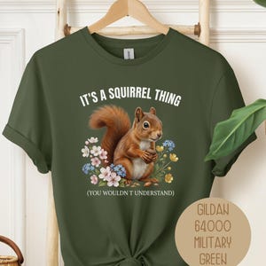 以下が含まれることがあります： ミリタリーグリーンの半袖Tシャツ。カラフルな花々に囲まれた茶色のリスのグラフィックが描かれています。上部には「IT'S A SQUIRREL THING」、下部には「(YOU WOULDN'T UNDERSTAND)」の文字があります。円形のラベルには「GILDAN 64000 MILITARY GREEN」と記載されています。