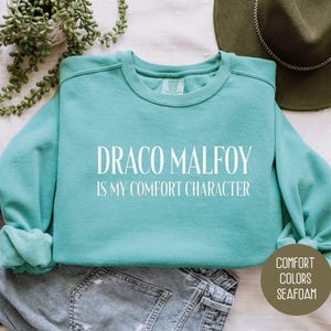 May include: Seafoam green crewneck sweatshirt with the text "DRACO MALFOY IS MY COMFORT CHARACTER" in white. The sweatshirt is folded on top of a pair of gray jeans. A green hat and a plant are in the background. The sweatshirt is part of the "Comfort Colors" collection.
