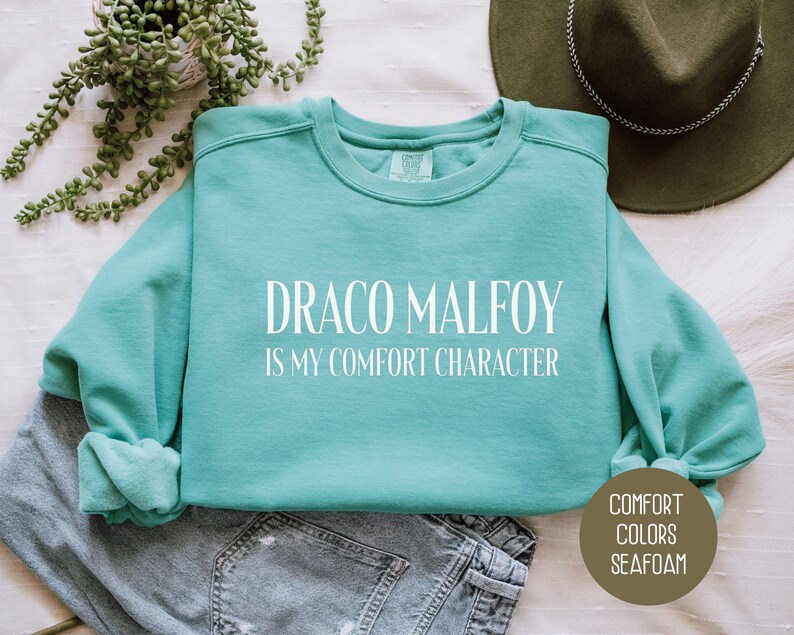 May include: Seafoam green crewneck sweatshirt with the text "DRACO MALFOY IS MY COMFORT CHARACTER" in white. The sweatshirt is folded on top of a pair of gray jeans. A green hat and a plant are in the background. The sweatshirt is part of the "Comfort Colors" collection.