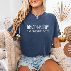 May include: A navy blue t-shirt with the text "DRACO MALFOY IS MY COMFORT CHARACTER" in white. The shirt is part of the "Comfort Colors Denim" collection. The model is wearing denim shorts and a cream-colored sweater.