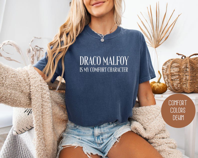 May include: A navy blue t-shirt with the text "DRACO MALFOY IS MY COMFORT CHARACTER" in white. The shirt is part of the "Comfort Colors Denim" collection. The model is wearing denim shorts and a cream-colored sweater.