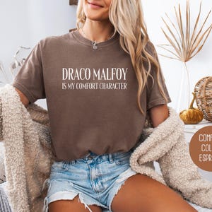 May include: Espresso-colored comfort colors t-shirt with the text "DRACO MALFOY IS MY COMFORT CHARACTER" in white. The shirt is worn with denim shorts and a cream-colored cardigan. The image also includes decorative elements.