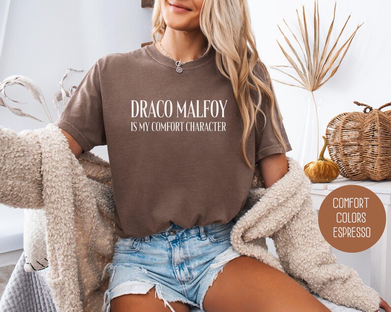 May include: Espresso-colored comfort colors t-shirt with the text "DRACO MALFOY IS MY COMFORT CHARACTER" in white. The shirt is worn with denim shorts and a cream-colored cardigan. The image also includes decorative elements.