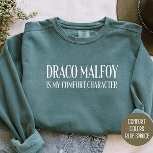 May include: Blue spruce-colored sweatshirt with the text "DRACO MALFOY IS MY COMFORT CHARACTER" in white. The sweatshirt is folded on top of a pair of blue jeans. The words "COMFORT COLORS BLUE SPRUCE" are printed on a brown circle.