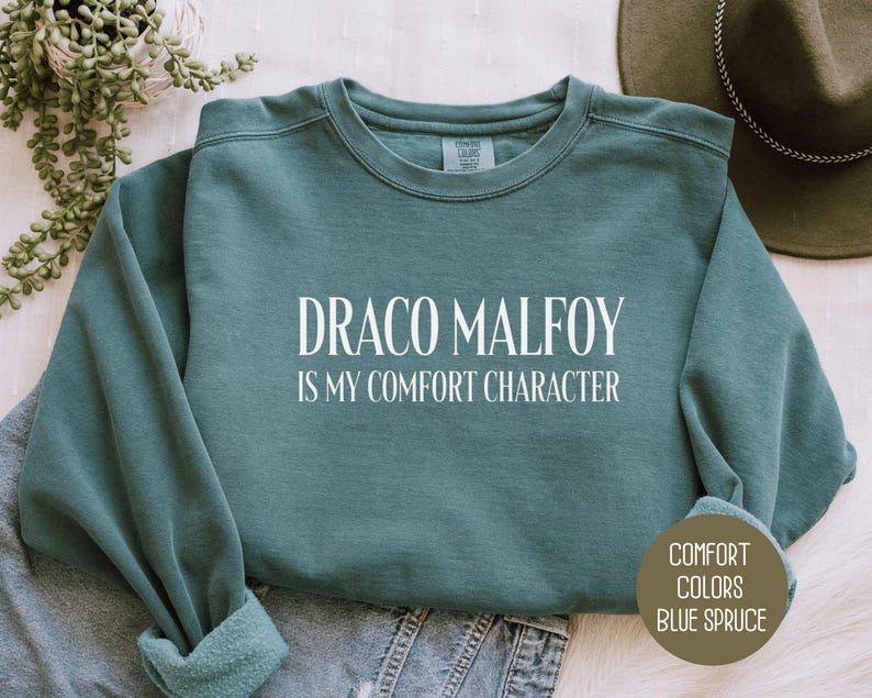 May include: Blue spruce-colored sweatshirt with the text "DRACO MALFOY IS MY COMFORT CHARACTER" in white. The sweatshirt is folded on top of a pair of blue jeans. The words "COMFORT COLORS BLUE SPRUCE" are printed on a brown circle.