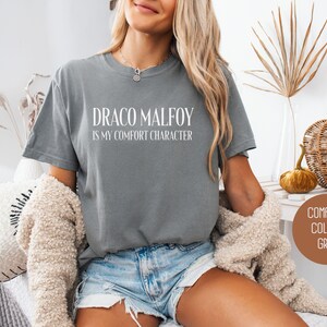 May include: Grey t-shirt with the text "DRACO MALFOY IS my comfort character" in white. The shirt is a comfort colors brand. The model is wearing denim shorts and a beige cardigan.