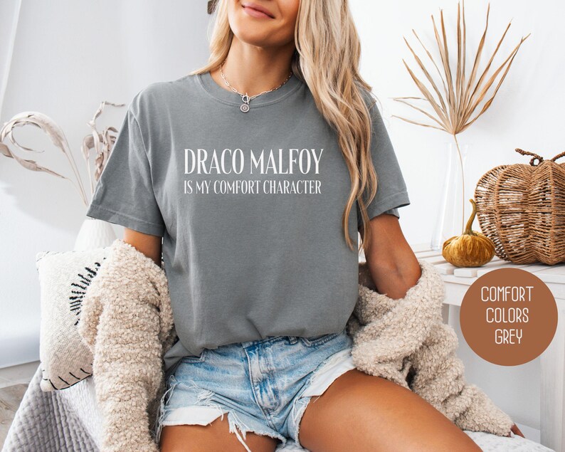 May include: Grey t-shirt with the text "DRACO MALFOY IS my comfort character" in white. The shirt is a comfort colors brand. The model is wearing denim shorts and a beige cardigan.