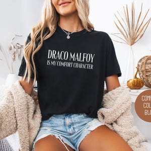 May include: Black t-shirt with the text "DRACO MALFOY IS MY COMFORT CHARACTER" in white. The shirt is a crew neck style. The image also includes a brown circle with the words "COMFORT COLORS BLACK".