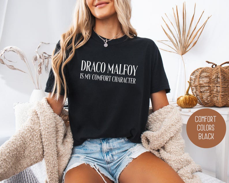 May include: Black t-shirt with the text "DRACO MALFOY IS MY COMFORT CHARACTER" in white. The shirt is a crew neck style. The image also includes a brown circle with the words "COMFORT COLORS BLACK".