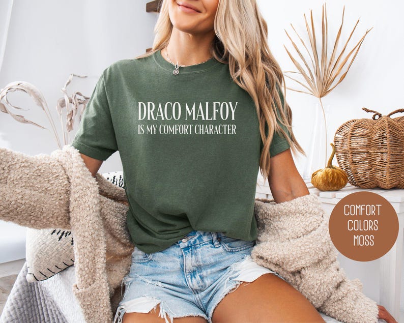 May include: Olive green t-shirt with the text "DRACO MALFOY is my comfort character" in white. The shirt is paired with denim shorts and a beige cardigan. A round sticker with "COMFORT COLORS MOSS" is visible.
