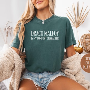 May include: Blue spruce t-shirt with the text "DRACO MALFOY is my comfort character" in white. The shirt is a solid color and has a crew neck. The image also shows a beige cardigan.