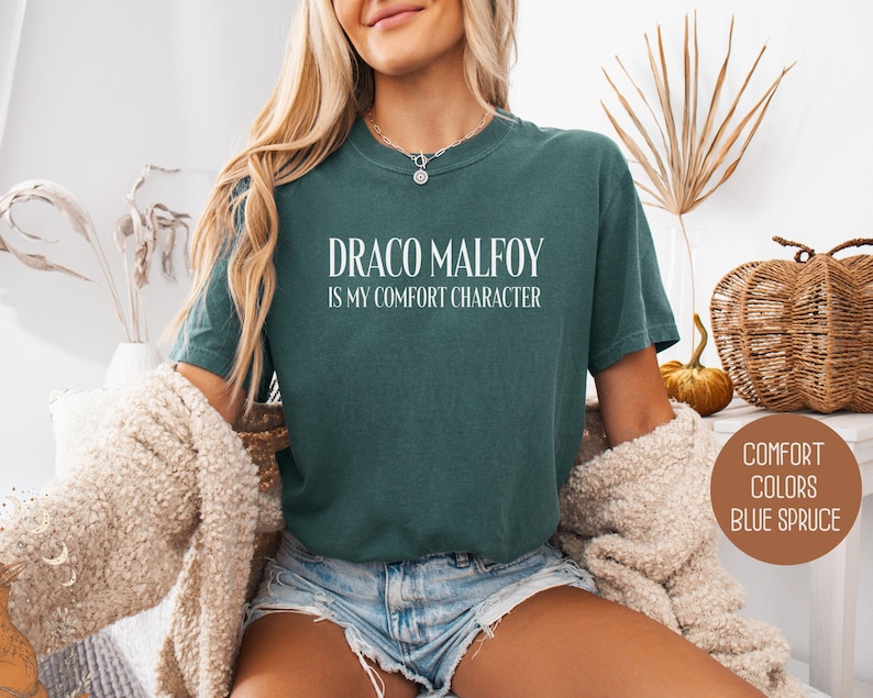 May include: Blue spruce t-shirt with the text "DRACO MALFOY is my comfort character" in white. The shirt is a solid color and has a crew neck. The image also shows a beige cardigan.