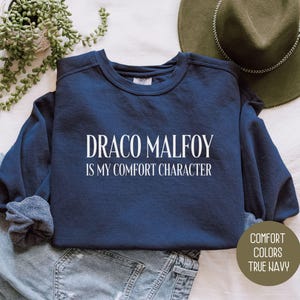 May include: A navy blue sweatshirt with the text "DRACO MALFOY IS MY COMFORT CHARACTER" in white. The sweatshirt is folded on top of a pair of blue jeans. A green hat and a plant are in the background.