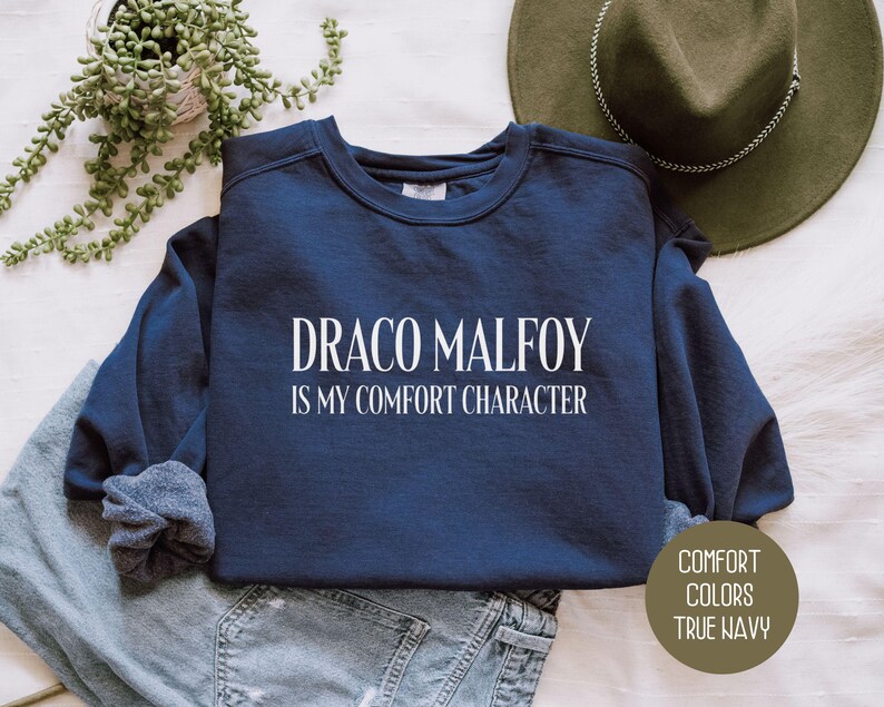 May include: A navy blue sweatshirt with the text "DRACO MALFOY IS MY COMFORT CHARACTER" in white. The sweatshirt is folded on top of a pair of blue jeans. A green hat and a plant are in the background.