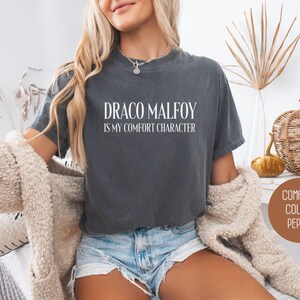 May include: A dark gray t-shirt with the text "DRACO MALFOY IS MY COMFORT CHARACTER" in white. The shirt is a comfort colors brand. The model is wearing denim shorts and a cream-colored cardigan.
