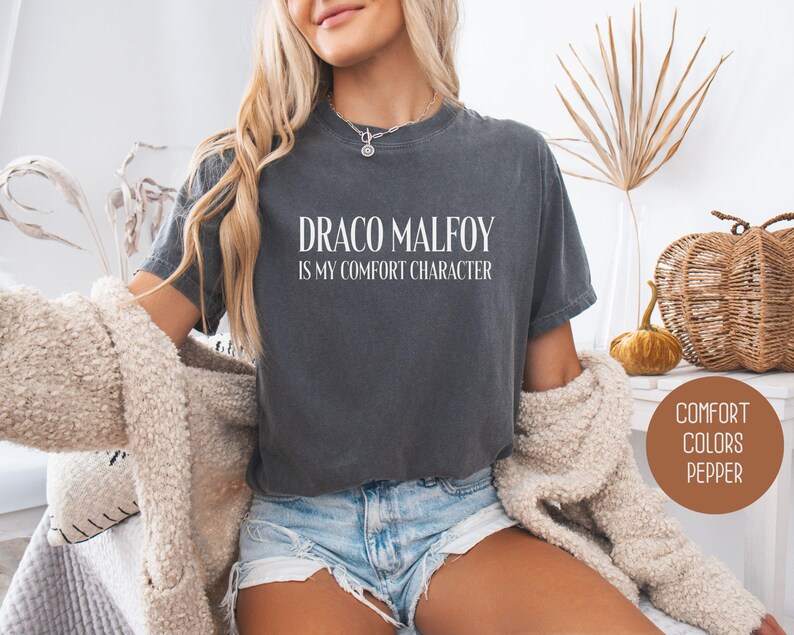 May include: A dark gray t-shirt with the text "DRACO MALFOY IS MY COMFORT CHARACTER" in white. The shirt is a comfort colors brand. The model is wearing denim shorts and a cream-colored cardigan.