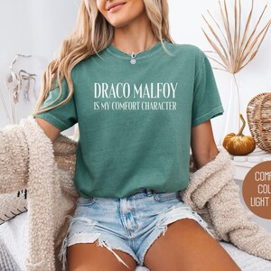 May include: Light green t-shirt with the text "DRACO MALFOY IS MY COMFORT CHARACTER" in white. The shirt is a comfort color and is light green. The person is wearing blue jean shorts and a cream-colored sweater.