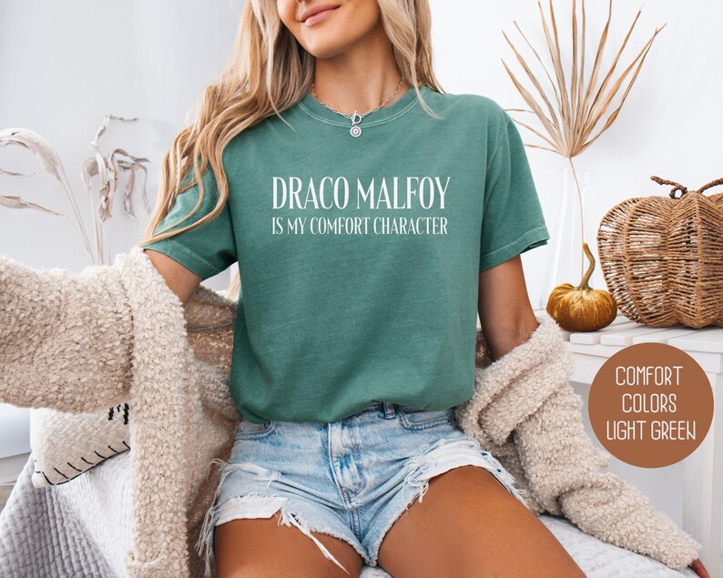 May include: Light green t-shirt with the text "DRACO MALFOY IS MY COMFORT CHARACTER" in white. The shirt is a comfort color and is light green. The person is wearing blue jean shorts and a cream-colored sweater.