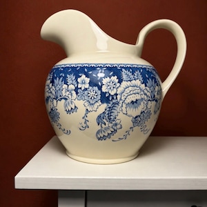 Può includere: Una brocca in ceramica color crema con corpo arrotondato, manico curvo e un ampio beccuccio. L'esterno è decorato con un motivo floreale blu che avvolge il centro, mettendo in risalto fiori e fogliame dettagliati.