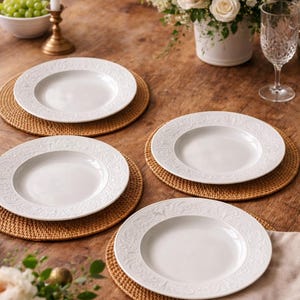 Peut inclure: Quatre assiettes en céramique blanche avec des motifs floraux en relief, chacune sur un dessous de plat rond en osier. Les assiettes sont disposées sur une table en bois, avec une bougie, des fleurs et un verre à vin.