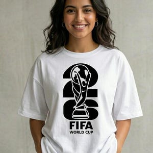 Peut inclure: Un t-shirt blanc à manches courtes arborant un grand motif graphique noir représentant le nombre 2026 avec la silhouette du trophée de la Coupe du Monde de la FIFA intégrée, au-dessus du texte FIFA WORLD CUP en lettres noires grasses.