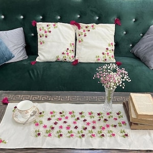 Op de afbeelding: Een witte rechthoekige tafelloper met geborduurde roze en paarse bloemen met groene ranken, vergezeld van twee bijpassende vierkante kussens met bloemenborduursel in de hoeken en roze kwastjes, een witte theekop en een stapel boeken.