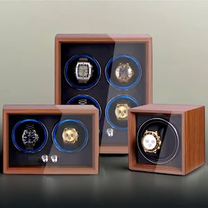 May include: Three wooden watch winders with glass tops. The winders are in different sizes and have a black interior with blue rotating cushions. The winders are on a gray surface.