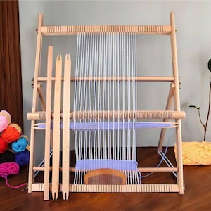 Op de afbeelding: Een houten weefgetouw met een lichtpaarse warp en een set houten weefgereedschap. Het weefgetouw is opgesteld op een houten standaard met een lichte houtkleur. Er zijn kleurrijke garenbollen op de voorgrond.