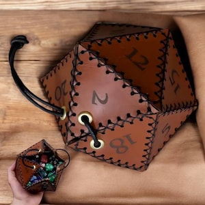 May include: A brown leather dice bag shaped like a twenty-sided die. The bag has a drawstring closure and is decorated with black stitching and gold eyelets. The numbers 1, 2, 5, 12, 18, and 20 are visible on the bag.